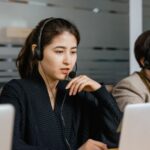Two people wearing headsets work at laptops in an office, focused on their screens.