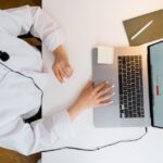 Person wearing headset works on a laptop at a desk with a notebook, pen, and coffee.