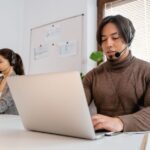 Man and woman wearing headsets working on laptops at desks in a bright office.