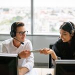 Two call center workers with headsets share a document while sitting at desks near large windows.