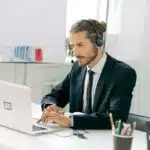 Man in suit with headset working on laptop at desk with pens, plants, and a whiteboard in the background.