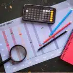 Revenue report chart with magnifying glass, calculator, colored pencils, and a pink notebook on a table.
