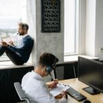 Two men wearing headsets work in an office; one sits by a window with a tablet, another at a desk.