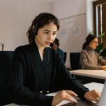 Woman with headphones works on laptop at a desk in an office with others in the background.