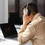 Person wearing headphones sits at a desk with a laptop, holding their head while looking at the screen.
