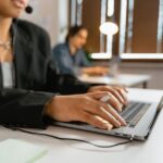 Person in a headset types on a laptop at a desk, with another person working in the background.