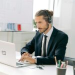 Man in a suit with a headset works on a laptop at a desk with office supplies and a whiteboard behind him.