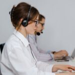 Two women wearing headsets work on laptops at a desk in an office setting.