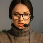 Woman wearing glasses and a headset speaks, working at a call center desk with another person in the background.