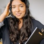Woman with long hair wearing a headset holds a clipboard and smiles at the camera in an office setting.