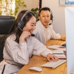 Two people in headsets sit at a desk, smiling and working on desktop computers in an office.