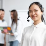 Woman with headset smiling, two people talking in the background holding a laptop in a bright office.