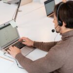Person wearing headset works on a laptop at a desk, with documents on a clipboard nearby.