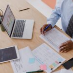 Man reviews documents at desk with laptop, tablet, phone, papers, and colorful sticky notes.