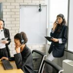 Three women in headsets work in a modern office; one types, two hold clipboards near windows and a whiteboard.