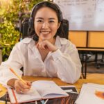 Woman in headset smiling at desk, writing in notebook with charts and keyboard nearby.