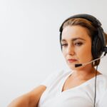 Woman wearing a white shirt and a headset with a microphone, sitting against a plain white background.