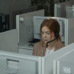 A woman with a headset sits at a desk in an office cubicle, looking at a computer monitor.