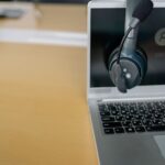 A headset with a microphone hangs on an open laptop displaying a login screen on a wooden desk.