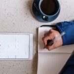 Person writing in a notebook next to a tablet displaying a calendar and a cup of coffee on a round table.