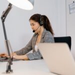 Woman wearing a headset works on a laptop at a desk in an office with another person nearby.
