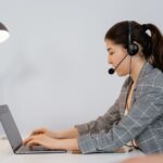 Woman in headset types on a laptop at a desk, working in an office with a lamp and plant nearby.