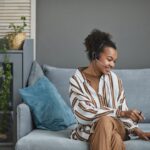 Woman wearing a headset smiles while working on a laptop, sitting on a gray couch with blue pillows.