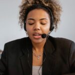 Woman with curly hair wearing a headset speaks at a desk, another person works in the background.