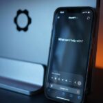 A smartphone displaying a ChatGPT prompt, “What can I help with?” rests against a laptop on a desk.