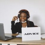 Woman wearing headphones smiles at laptop, sitting at desk with a sign that reads “ADVICE.”.